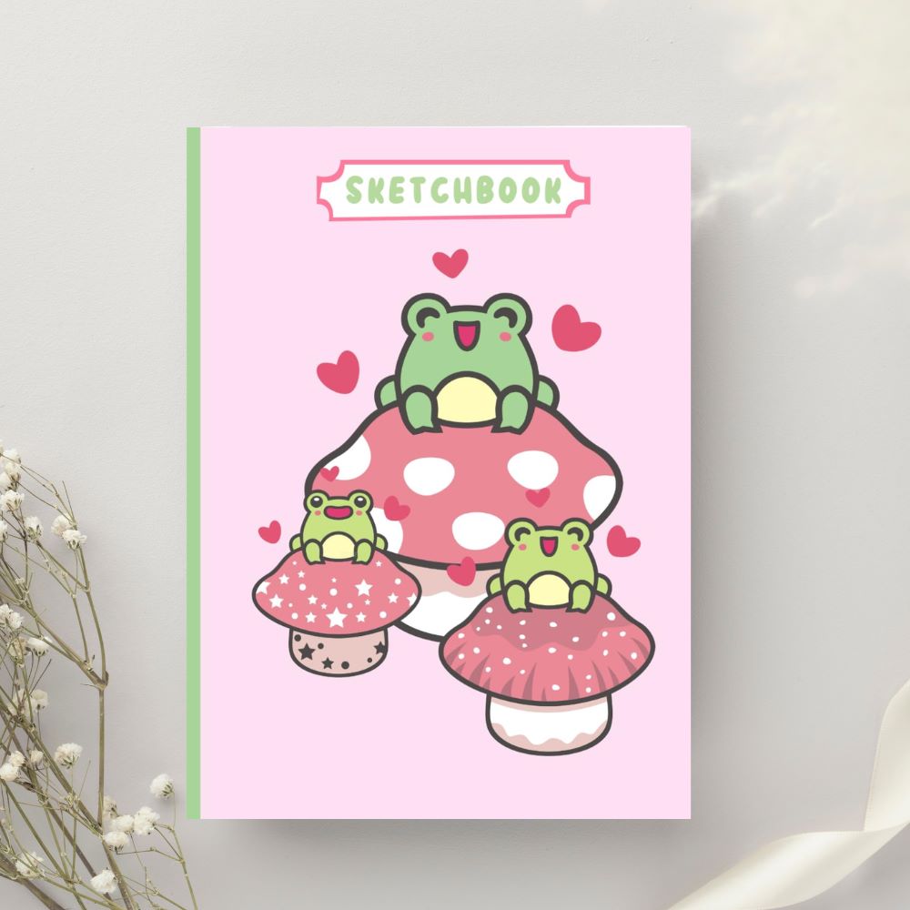 Children's sketchbook with a playful cover design featuring cartoon frogs on colorful mushrooms, set against a soft pink background with decorative hearts, displayed next to white flowers on a light grey wall.