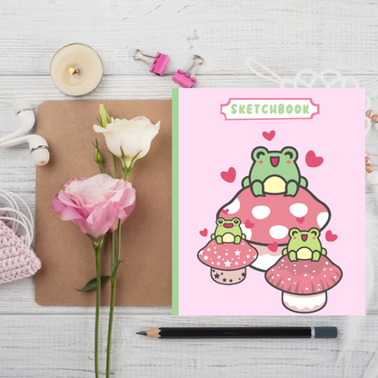 Artistic sketchbook cover featuring cute cartoon frogs on colorful mushrooms, styled on a wooden surface with pink and white roses, a candle, headphones, and a pencil, ideal for creative children's stationery.