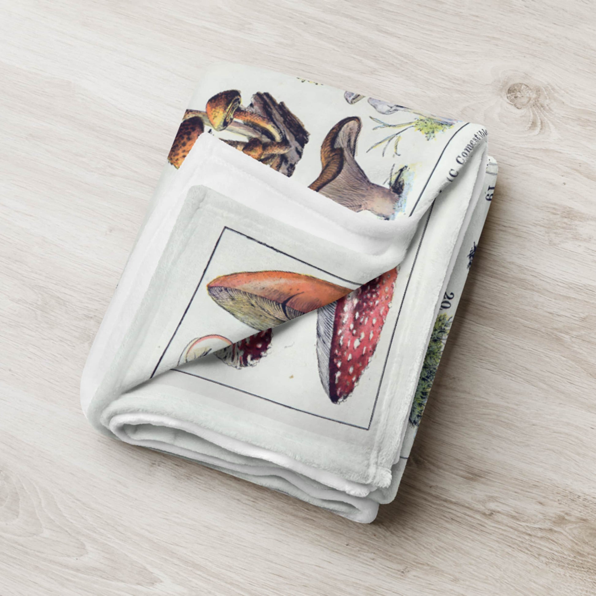 Soft fleece blanket folded neatly on a wooden floor, showcasing a vintage print of mushrooms in rich, earthy tones, a cozy addition to any home with a touch of botanical charm