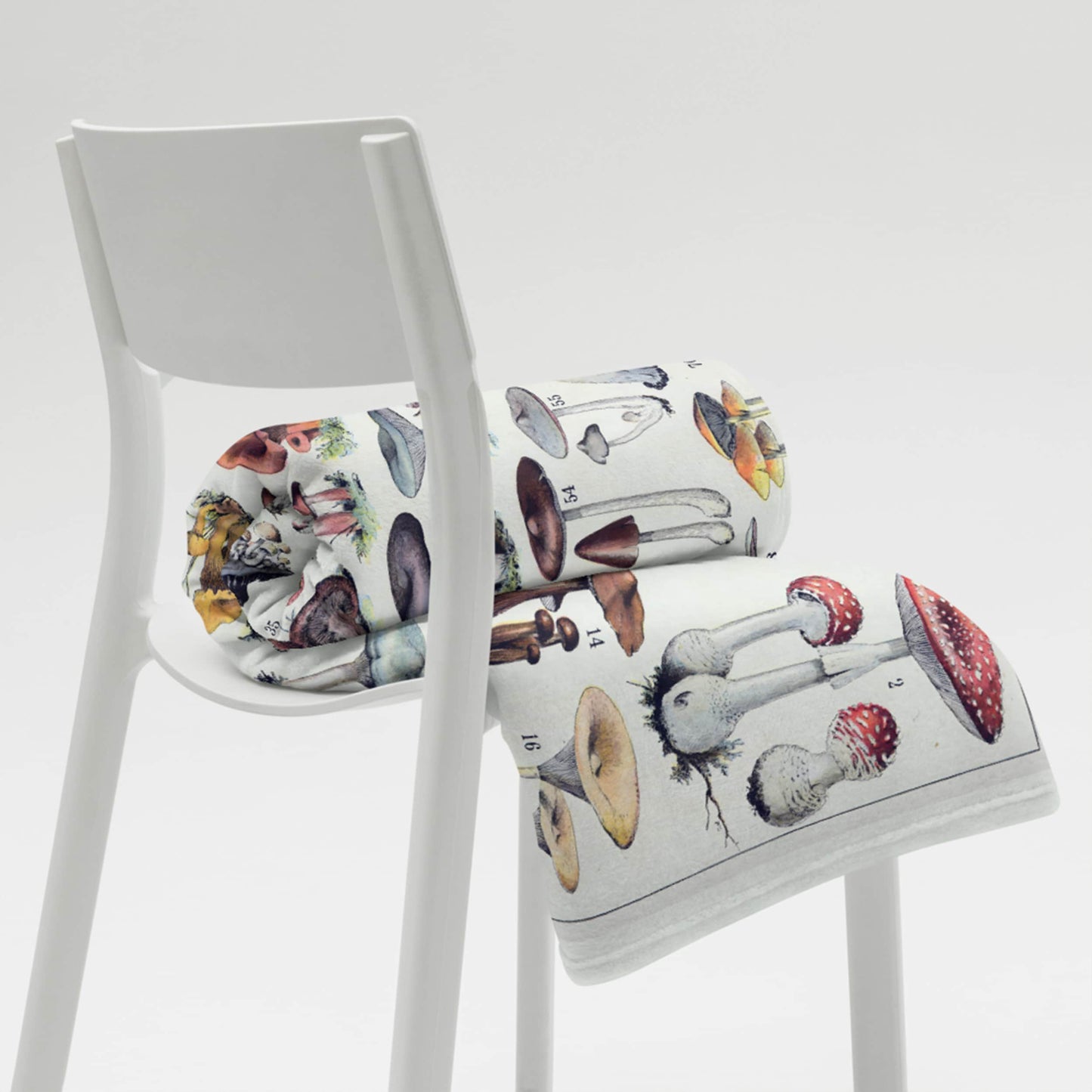 A modern white chair presenting a rolled-up fleece blanket with a detailed, colorful print of various mushrooms, juxtaposing contemporary furniture design with naturalistic elements