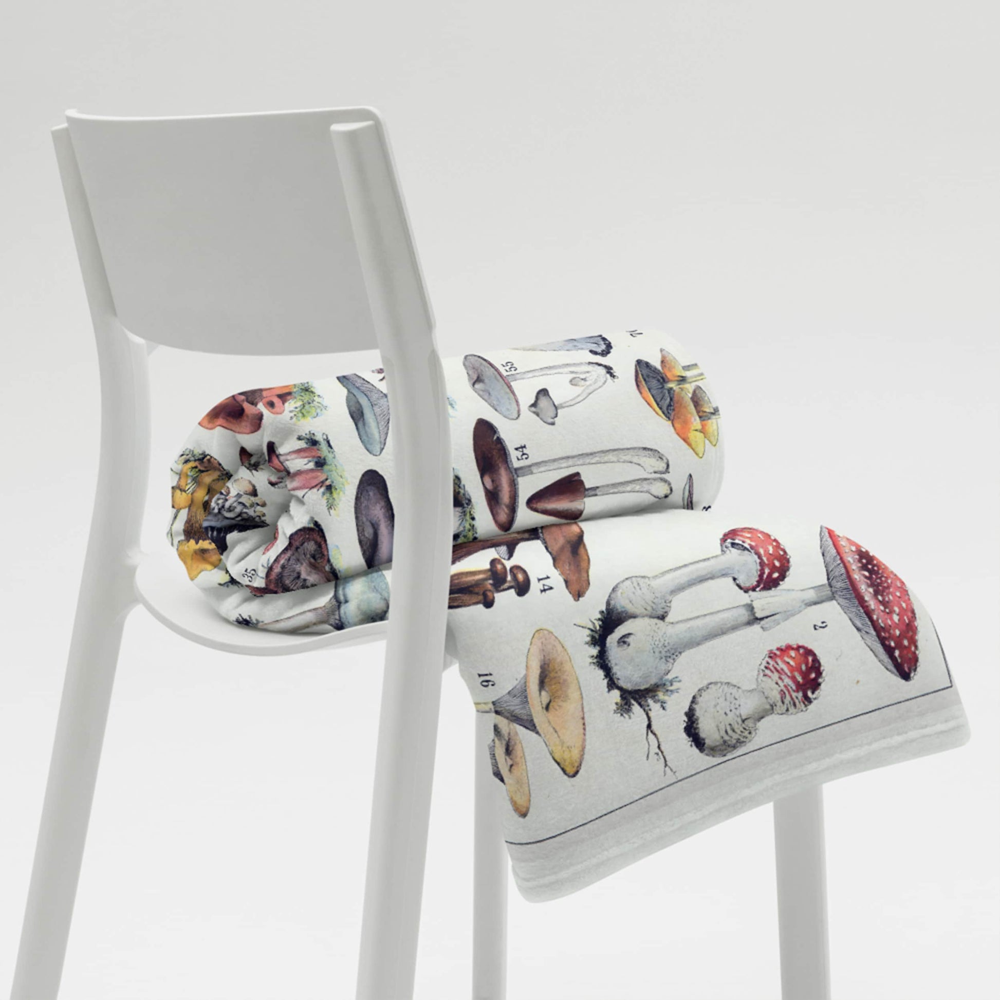 A modern white chair presenting a rolled-up fleece blanket with a detailed, colorful print of various mushrooms, juxtaposing contemporary furniture design with naturalistic elements